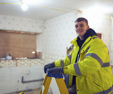 A construction student in a yellow jacket standing on a ladder, smiling at the camera.