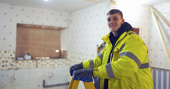 A construction student in a yellow jacket standing on a ladder, smiling at the camera.