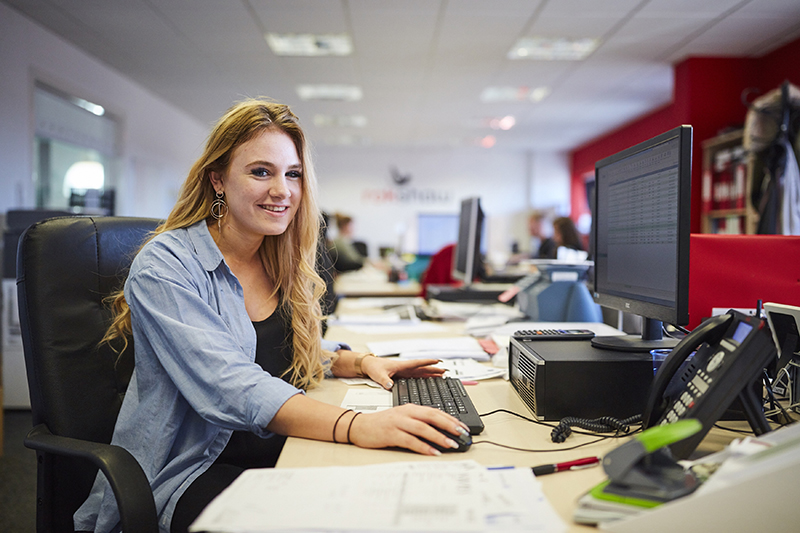 In an office, a woman apprentice sits at a desk, smiling and looking towards the camera.