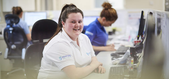 In an office, a woman apprentice sits at a desk, smiling and looking towards the camera.