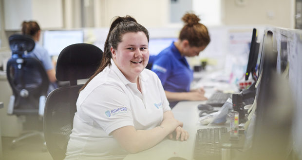 In an office, a woman apprentice sits at a desk, smiling and looking towards the camera.