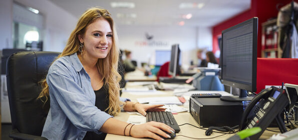 In an office, a woman apprentice sits at a desk, smiling and looking towards the camera.